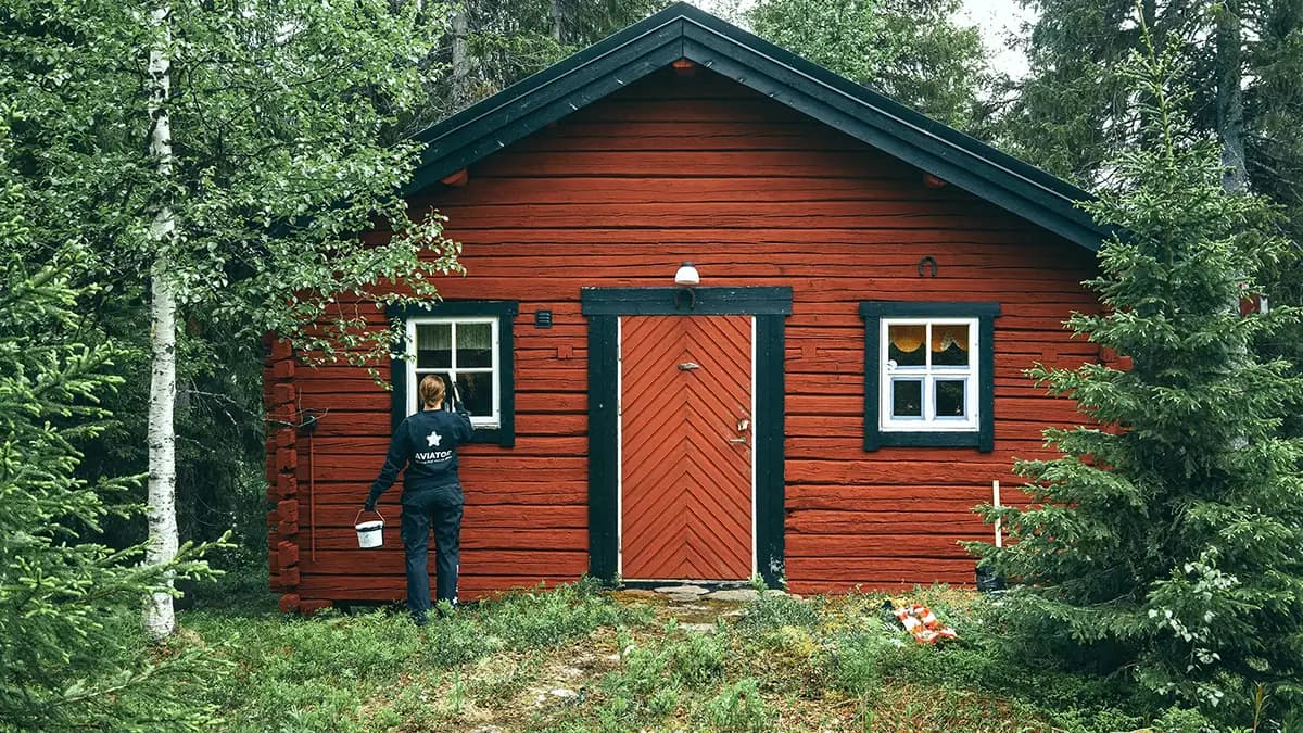En person som holder en bøtte står foran et rødt hus omgitt av gress og trær.